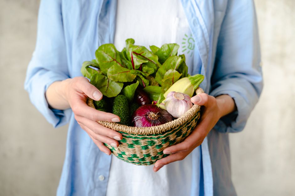 A person wearing a light blue shirt holding a woven basket filled with fresh garden vegetables, including leafy greens, a cucumber, a garlic bulb, and a red onion. The background is a neutral, slightl